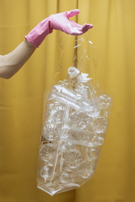 A person wearing a pink rubber glove is holding a large, transparent plastic bag filled with crushed and empty plastic bottles. The bottles are clear and of various sizes, with some caps visible among the debris. The background consists of a plain yellow curtain or panel, emphasizing the focus on the plastic waste. The scene suggests an act of plastic waste collection or disposal, aligning with practices of private rubbish removal services that handle environmentally conscious waste segregation, often involving on-site clearance or independent collection methods. The image highlights the importance of proper disposal of plastic materials, which waste management companies such as Rubbish Clearance Crystal Palace often assist with, supporting sustainable waste handling solutions in the local area.