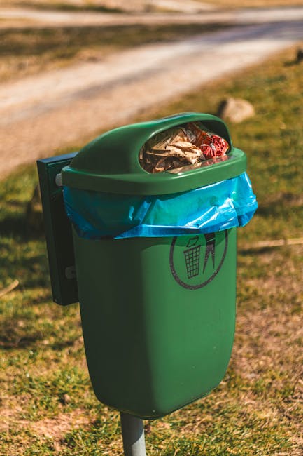 A green outdoor trash bin with a rounded, hinged lid is positioned on a grassy area beside a concrete pathway. The lid is partially open, revealing assorted waste including paper, plastic wrappers, and foil. A blue plastic bag lines the top of the bin, secured around the opening, with a black printed symbol of a waste bin and a person throwing rubbish. The surrounding environment includes patches of grass, some rocks, and a blurred background of a dirt path or road with natural, warm lighting suggesting daytime. The scene reflects an example of private waste collection and regular rubbish disposal, supporting eco-friendly rubbish removal services as offered by companies such as rubbishclearancecrystalpalace.com for alternative rubbish handling in outdoor public spaces.
