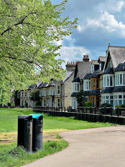 In a residential area, a paved pathway runs alongside a grassy verge bordered by a black wooden fence, with a large deciduous tree on the left side that has vibrant green leaves and spreading branches. Behind the fence, a row of Victorian-style terraced houses with multiple stories, bay windows, and slate roofs is visible, featuring brick facades in shades of beige and grey, some with contrasting window surrounds. Two black waste bins with rounded tops and blue lids are positioned at the edge of the pathway, partially surrounded by the grass and pavement. The scene is illuminated by natural daylight, with a partly cloudy sky overhead, indicating a bright and clear day. The image, associated with solutions for private waste handling and rubbish removal, subtly reflects the environment in which independent collection services might operate outside of local authority disposal channels, illustrating the typical setting for rubbish clearance or house clearance projects in a suburban context.