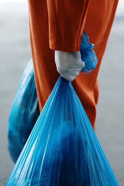 Close-up of a person wearing a white protective glove, holding the blue handle of a large blue plastic rubbish bag filled with waste. The individual is dressed in an orange high-visibility coverall, suggesting a work-related environment such as waste collection or rubbish removal. The background is blurred but appears to be an outdoor setting, possibly on a paved area or driveway. The scene highlights the process of waste handling, with the bag partially visible and bulging with waste material, indicating recent collection or preparation for disposal. The overall lighting is natural, emphasizing clean and practical attire suitable for professional rubbish clearance services. This image exemplifies the kind of independent waste collection or on-site rubbish removal activity that might be undertaken by companies like [COMPANY_NAME] specializing in domestic or commercial refuse clearance.