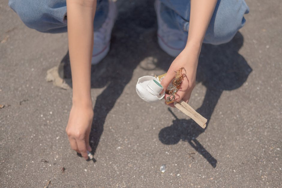 A person wearing blue jeans and white sneakers is crouched on a paved outdoor surface, holding a small collection of debris and rubbish in their right hand. The debris includes a torn, white, plastic disposable cup with some foam material still attached, as well as small pieces of wood and tangled string or plastic fibers. Their left hand rests near the ground, close to a few tiny scattered fragments. The scene appears to be in an open area, possibly a driveway or paved yard, with no other objects visible in the background. The lighting suggests natural daylight, highlighting the texture of the gritty asphalt and the details of the rubbish, emphasizing the importance of rubbish removal efforts. This image potentially relates to the context of waste collection or public clean-up activities, as carried out by professional rubbish clearance services such as those provided by rubbishclearancecrystalpalace.com, especially in residential or community outdoor spaces in the Crystal Palace area.