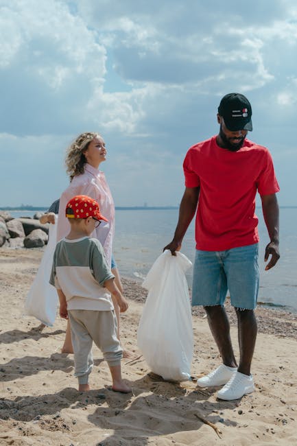 A group of three individuals, including a man and two children, are participating in a beach clean-up activity along the shoreline. The man, dressed in a red T-shirt, light blue denim shorts, and a black cap, is holding a white plastic bag in his right hand, appearing to be collecting litter from the sand. To his left, a young boy wearing a grey T-shirt, beige shorts, and a red cap with a pattern, stands barefoot on the sandy beach, holding a similar white bag. Behind the children, a woman with curly blonde hair, wearing a light pink long-sleeved top, observes the scene, standing slightly further back near the water's edge. The background features a partly cloudy sky with bright sunlight shining on the beach, stones piled along the shoreline, and calm water stretching towards the horizon. This scene reflects an outdoor waste removal effort, emphasizing personal initiatives to manage rubbish outside of official waste collection services, with the focus on collecting litter in a natural environment to support cleanliness and environmental conservation.